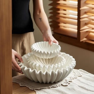 Könnte beinhalten: Ein Set aus drei weißen Keramikschalen mit gewelltem Rand. Die Schalen sind ineinander gestapelt, wobei die größte Schale unten und die kleinste oben liegt. Die Schalen stehen auf einer neutralfarbenen Oberfläche.