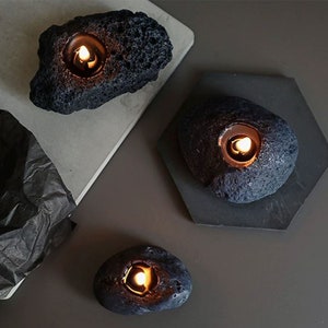 May include: Three black lava rock candle holders with a small, round, white candle burning inside each one. The candle holders are on a gray surface.