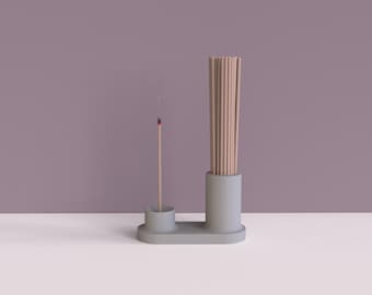 May include: A gray ceramic incense holder with two compartments. One compartment holds a single incense stick, while the other holds a bundle of incense sticks. The incense holder is on a white surface.