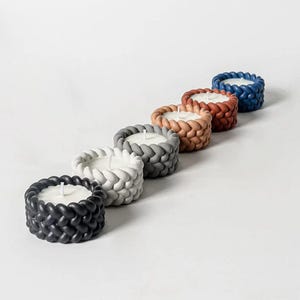 May include: A set of six braided candle holders in various colors, including black, gray, white, brown, and red. The candle holders are arranged in a row, with the black candle holder at the front and the red candle holder at the back.