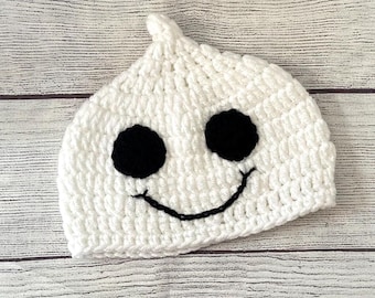 May include: A white crocheted hat shaped like a ghost, featuring two large black eyes and a black stitched smile. The hat is handmade and sits on a light-colored wooden surface.