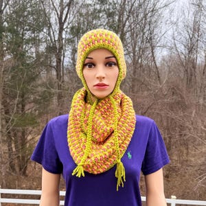 May include: A handmade crochet hat and cowl set in shades of yellow, pink, and green. The hat is a close-fitting balaclava style, and the cowl is a wide loop. The set is displayed on a mannequin, showcasing the textured stitch pattern.