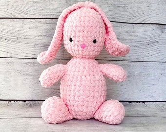 Könnte beinhalten: Ein rosa gehäkeltes Plüsch-Hasenspielzeug mit schwarzen Augen und einer rosa Nase. Der Hase sitzt auf einer weißen Holzoberfläche.