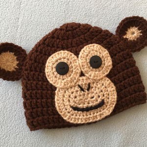 May include: Brown crocheted monkey hat with beige crocheted ears and face. The hat has black button eyes and a black crocheted mouth.