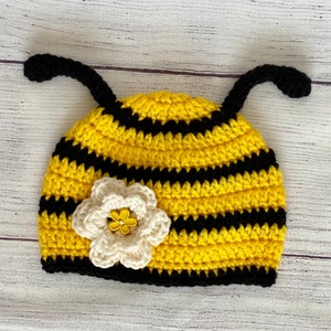 Può includere: Un cappello da ape in crochet giallo e nero con un fiore bianco in crochet sulla cima. Il cappello ha due antenne in crochet nere.
