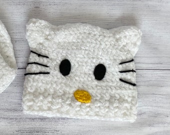 Puede incluir: Sombrero blanco de crochet con diseño de cara de gato. El sombrero tiene bigotes, ojos y una nariz amarilla bordados en negro.
