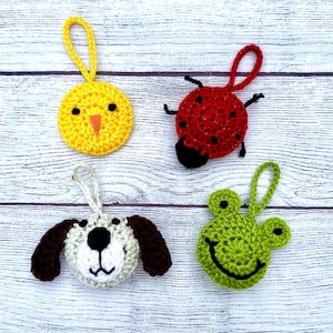 May include: Four crocheted animal ornaments, a yellow chick, a red ladybug, a brown and white dog, and a green frog, are displayed on a white wooden surface.
