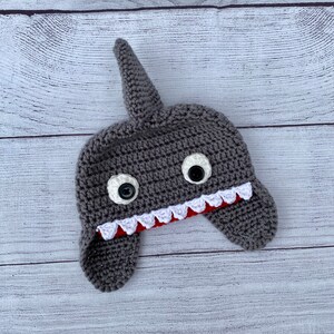 May include: Gray crocheted shark hat with white eyes and red and white teeth. The hat has ear flaps.