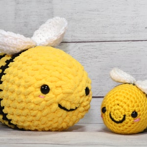 May include: Two crocheted bee plushies. The larger bee is yellow with black stripes and white wings. The smaller bee has the same design. Both have smiling faces with black eyes and pink cheeks. The background is a light wood.