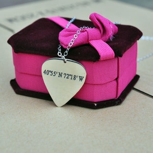May include: A silver-colored pendant necklace with a guitar pick shape, engraved with coordinates. The pendant hangs from a delicate chain and rests on a pink ribbon-wrapped box with a dark brown velvet top. The box is tied with a pink bow.