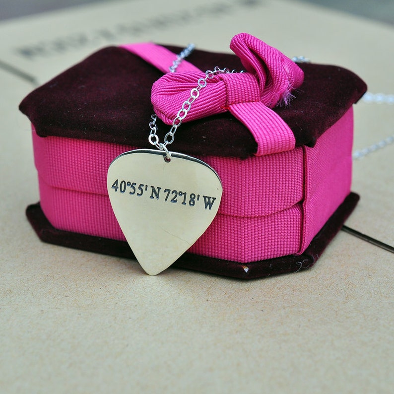 May include: A silver-colored pendant necklace with a guitar pick shape, engraved with coordinates. The pendant hangs from a delicate chain and rests on a pink ribbon-wrapped box with a dark brown velvet top. The box is tied with a pink bow.