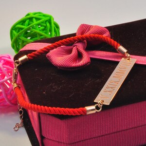 May include: A red string bracelet with a gold bar charm engraved with the Roman numerals "V.XXX.MMVII". The bracelet is displayed on a pink velvet box with a red bow.
