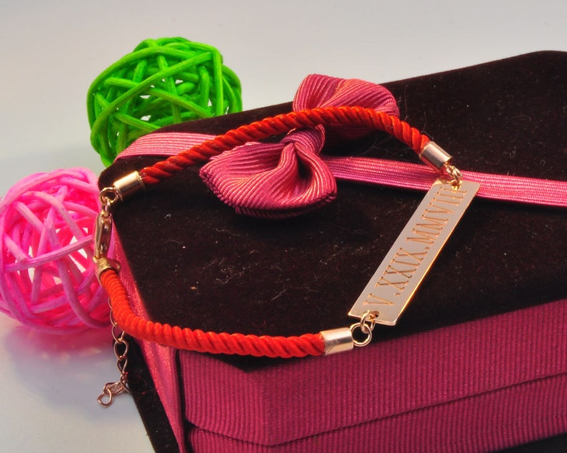 May include: A red string bracelet with a gold bar charm engraved with the Roman numerals "V.XXX.MMVII". The bracelet is displayed on a pink velvet box with a red bow.