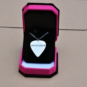May include: A silver guitar pick pendant necklace with the coordinates 40&deg;55' N 72&deg;18' W engraved on it. The pendant is displayed in an open black jewelry box with a pink and black exterior.
