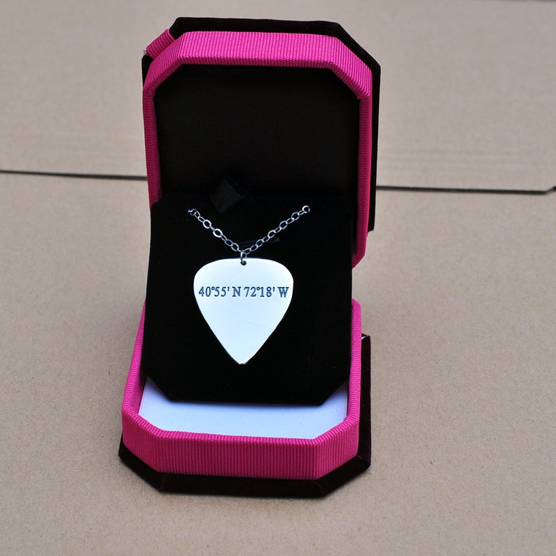 May include: A silver guitar pick pendant necklace with the coordinates 40&deg;55' N 72&deg;18' W engraved on it. The pendant is displayed in an open black jewelry box with a pink and black exterior.