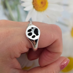 May include: A silver ring with a black paw print design. The ring is on a finger.