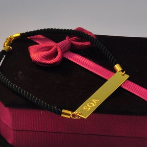 May include: A black cord bracelet with a gold bar charm engraved with the letters "SOA". The bracelet is displayed in a red velvet box with a pink ribbon.