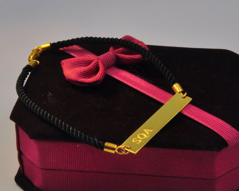 May include: A black cord bracelet with a gold bar charm engraved with the letters "SOA". The bracelet is displayed in a red velvet box with a pink ribbon.