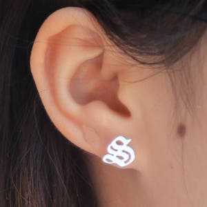 May include: A close-up of a silver stud earring in the shape of a stylized letter "S" is shown on an ear. The earring is small and simple, with a smooth, reflective surface. The ear is visible in the image.