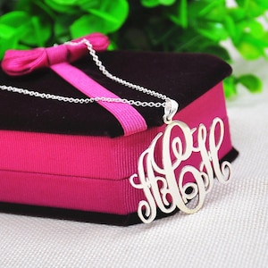 May include: A silver necklace with a monogram pendant in a pink velvet gift box. The pendant is a large, cursive script monogram with three letters.