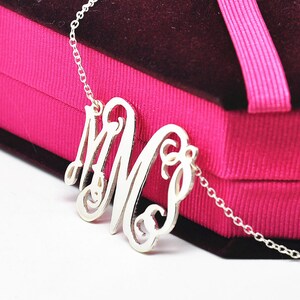 May include: A silver-colored monogram necklace featuring the intertwined letters "M" and "S". The pendant hangs from a delicate chain, presented against a pink gift box with a dark burgundy interior and a pink ribbon.