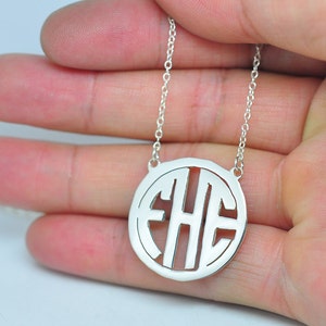 May include: A silver-colored monogram necklace featuring the letters EHE within a circular pendant. The pendant hangs from a delicate chain. The necklace is held in a person's hand.