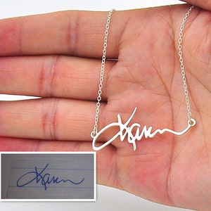 Pode incluir: Um colar prateado com um pendente com nome em estilo manuscrito. O nome em it&aacute;lico &eacute; exibido em uma m&atilde;o. Uma imagem menor mostra a assinatura no papel.