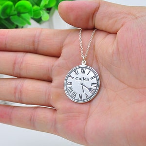 May include: A silver pendant necklace with a clock face design. The clock face has Roman numerals and the name "Cullen" printed on it. The clock hands indicate approximately 5:27. The necklace is held in a person's hand.