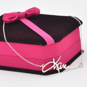 Pode incluir: Um colar prateado com o nome "Xam" em fonte cursiva. O colar &eacute; exibido em uma caixa de presente rosa com uma parte superior marrom escuro e um la&ccedil;o rosa.