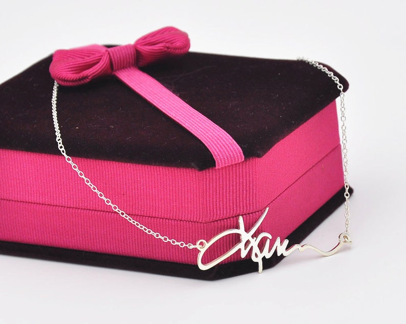 Pode incluir: Um colar prateado com o nome "Xam" em fonte cursiva. O colar &eacute; exibido em uma caixa de presente rosa com uma parte superior marrom escuro e um la&ccedil;o rosa.