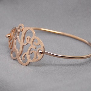 May include: A rose gold bangle bracelet with a delicate, openwork design featuring a monogram. The bracelet is made of metal and has a polished finish.