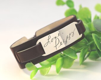May include: A dark brown leather wrap bracelet with a silver rectangular plate. The plate features handwritten text that reads "love, Peggy". The bracelet is wrapped around a green leafy plant.