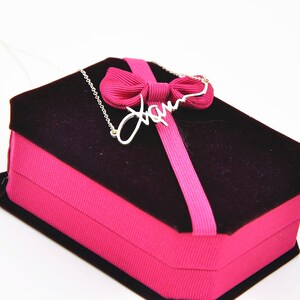 Pode incluir: Um colar prateado com o nome "Jam" em fonte cursiva, repousando sobre uma caixa de joias de veludo preto com uma fita e la&ccedil;o rosa. A caixa &eacute; retangular e tem uma base rosa.