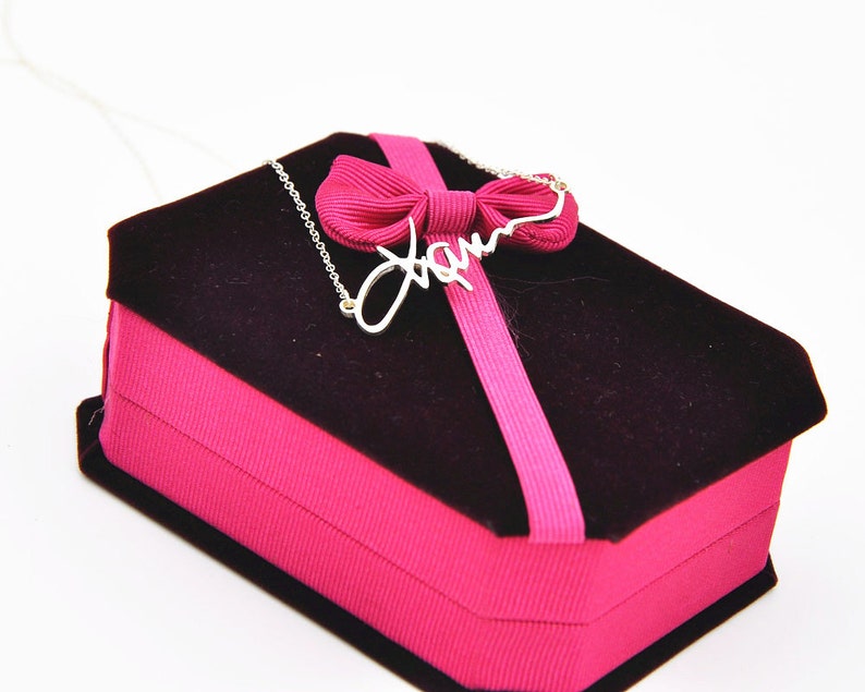 Pode incluir: Um colar prateado com o nome "Jam" em fonte cursiva, repousando sobre uma caixa de joias de veludo preto com uma fita e la&ccedil;o rosa. A caixa &eacute; retangular e tem uma base rosa.