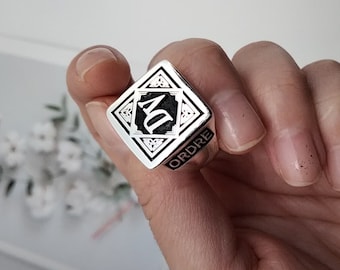 May include: A silver ring with a black square design featuring the letters "AD" inside a Celtic knot pattern. The ring is inscribed with the word "ORDRE".