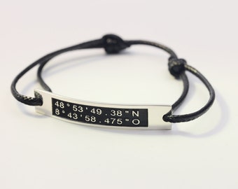 May include: Black cord bracelet with a silver rectangular tag engraved with the coordinates "48° 53' 49 . 38" N 8° 43' 58 . 475" O"