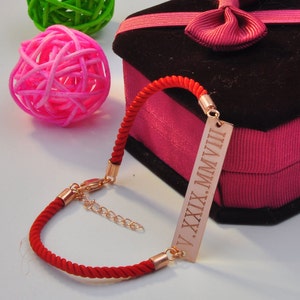 May include: Red string bracelet with a gold bar charm engraved with the Roman numerals "V.XXIX.MMVIII".