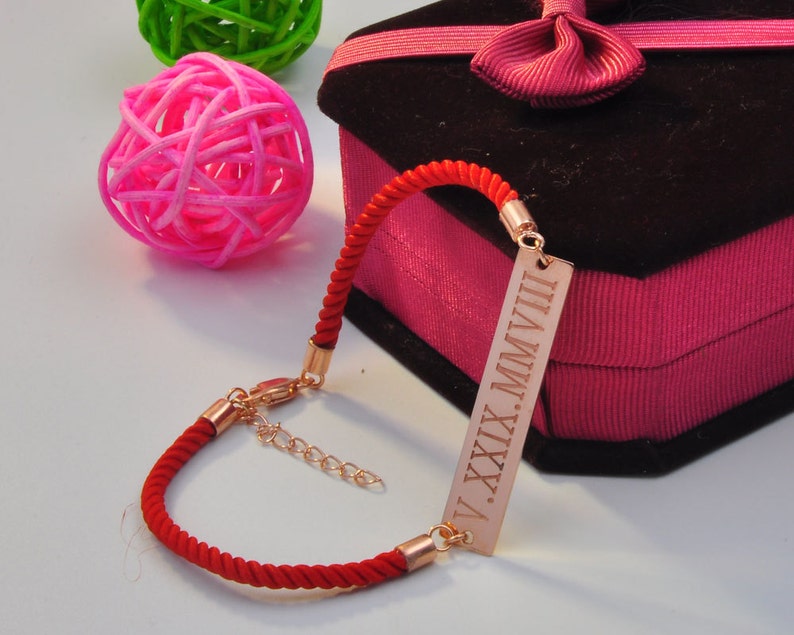May include: Red string bracelet with a gold bar charm engraved with the Roman numerals "V.XXIX.MMVIII".