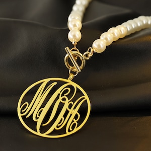 Peut inclure: Un collier en or avec un grand pendentif rond orné d'un monogramme. Le pendentif est en or et présente une écriture cursive délicate. Le collier est composé de perles blanches et possède un fermoir en or.