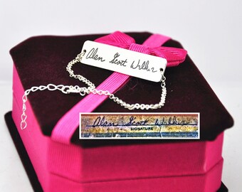 May include: A silver bracelet with a tag engraved with the name "Alan Scott Wilker" in cursive. The bracelet is presented on a pink gift box with a magenta ribbon.