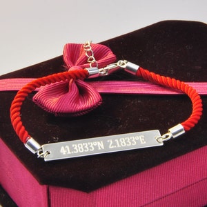 May include: A red rope bracelet with a silver plate engraved with the coordinates "41.3833&deg;N 2.1833&deg;E". The bracelet is displayed in a red velvet box with a pink ribbon.