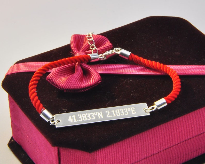 May include: A red rope bracelet with a silver plate engraved with the coordinates "41.3833&deg;N 2.1833&deg;E". The bracelet is displayed in a red velvet box with a pink ribbon.