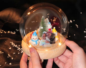 Puede incluir: Un globo de nieve de cristal transparente con una base de madera, que presenta una escena navideña en miniatura. En el interior, hay un pequeño árbol de Navidad, muñecos de nieve y una foto enmarcada. La base tiene pequeñas luces.