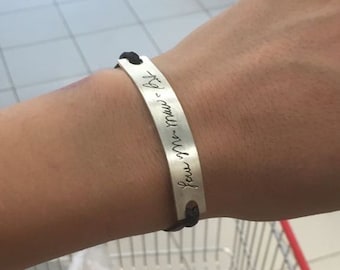 May include: Silver bracelet with black cord. The bracelet features engraved cursive text. The bracelet is worn on a wrist.