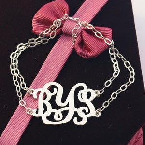 Op de afbeelding: Een zilverkleurige armband met de initialen "BYS" in sierlijke letters. De armband bestaat uit twee fijne kettingen. Het wordt gepresenteerd op een donkerrode ondergrond met een roze lint.