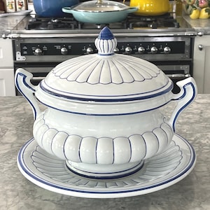 May include: A white ceramic tureen with a matching lid and base plate, featuring blue painted trim and vertical fluted detailing. The tureen has two curved handles on the sides and a pointed finial on the lid, all decorated with blue lines.