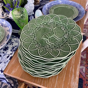 May include: A stack of green ceramic plates with a raised deer and grape design. The plates have a scalloped edge and are part of a larger table setting with other decorative items, including glassware and floral arrangements.