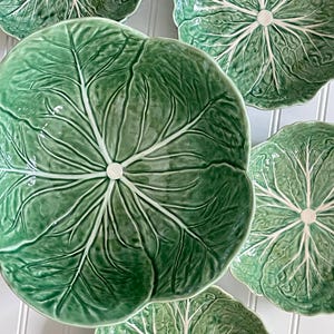 May include: A collection of five ceramic bowls shaped like green cabbage leaves. Each bowl features a glossy finish with realistic embossed leaf veins radiating from a central white circular point, mimicking the natural texture of a cabbage.