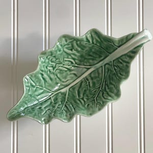 May include: A ceramic dish shaped like a green leaf with a glossy finish. The dish features a detailed embossed vein pattern across the surface and a prominent stem extending from one end, mimicking the natural appearance of a real plant leaf.
