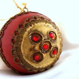 Puede incluir: Un adorno navideño rojo con ribetes dorados y joyas rojas. El adorno tiene un diseño tradicional.